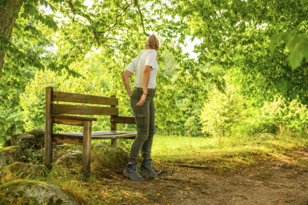 Young woman in summer clothes standing next to a wooden bench in sunny forest, Linnés Råshult,
