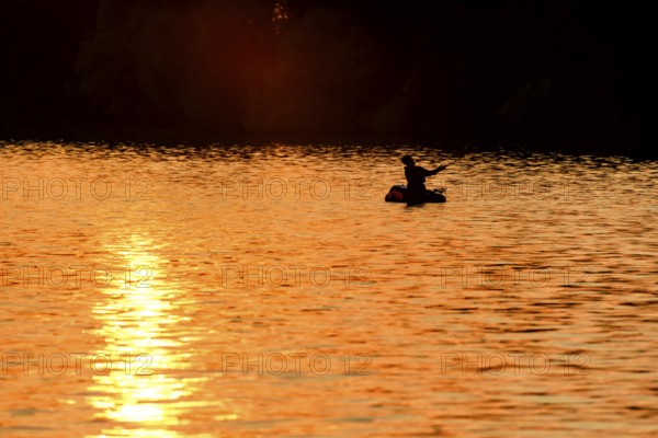 Angler sport fisherman in his belly boat in orange-red lake at sunset, silhouette in calm water,