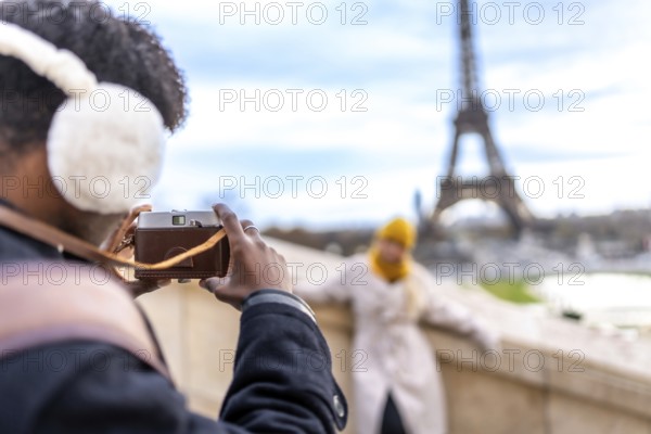 Young man wearing earmuffs and using a vintage camera to photograph his female partner posing with