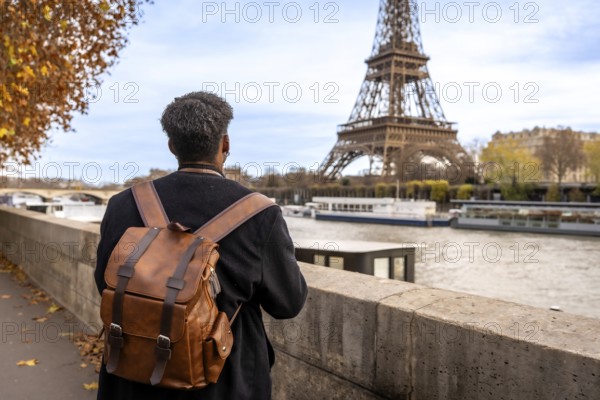 Person with travel backpack standing by the seine, gazing at the eiffel tower amid autumn foliage