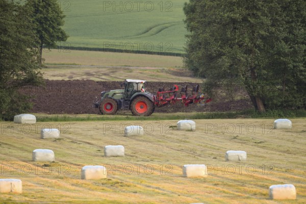 A tractor plows a field in a rural landscape with round bales of hay bales in the foreground,