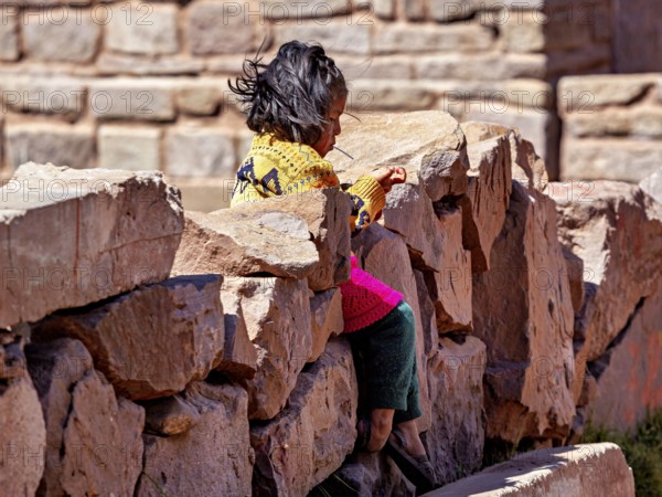 Little girl in colorful dress sitting on a stone wall and smiling, children from Taquile Island in