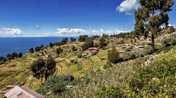 Village with huts on a hill surrounded by green fields and a blue lake under clear skies, the