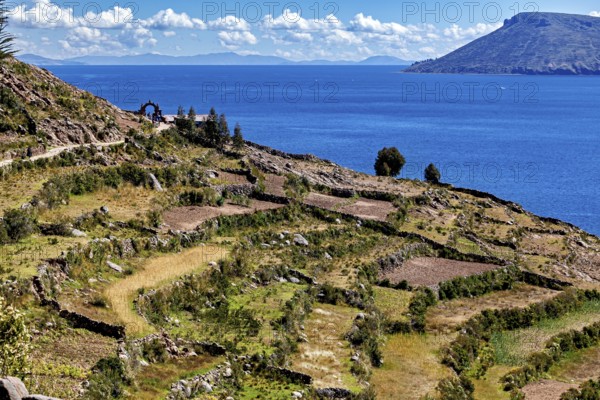 Terraced fields stretch down to a clear blue lake, framed by stone arches and hills, the