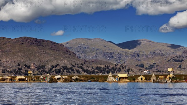 Reed huts on the shores of a tranquil lake in front of a vast mountain landscape under a blue sky,