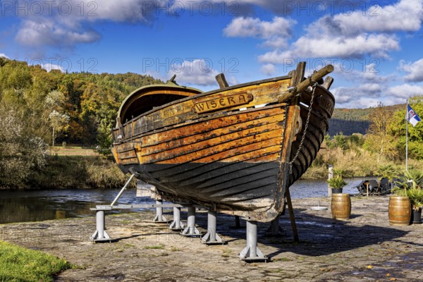 Historic wooden boat on riverbank, surrounded by autumn trees under a clear sky, the port of