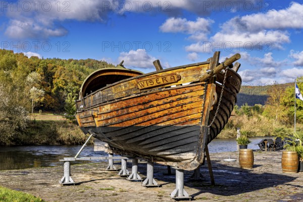 Historic wooden boat on the riverbank on supports, surrounded by autumn trees under a blue sky, the