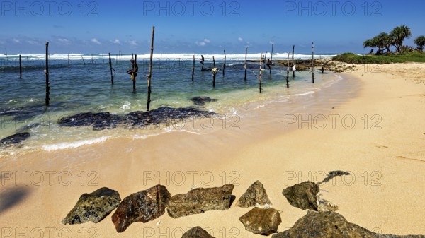 Quiet beach with clear waves and wooden poles under blue skies, Koggala beach with stilt fishermen