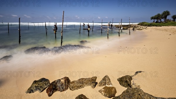 Clear beach with visible rocks and wooden posts in the water, Koggala beach with stilt fishermen in