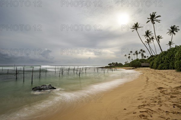 Tropical beach with soft waves, palm trees and cloudy sky, Koggala beach with stilt fishermen in