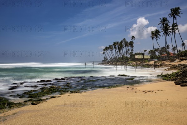 A sun-drenched beach with palm trees and waves hitting the rocks, Koggala beach with stilt