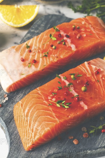 Two pieces of salmon fillet sit on a wooden cutting board. They are topped with herbs and pink