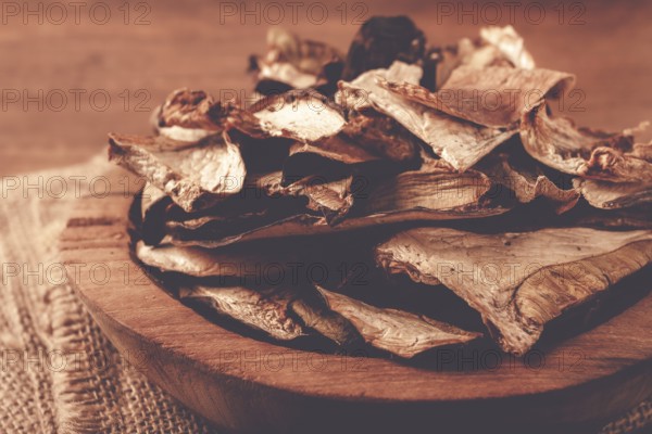 A collection of dried mushrooms fills a wooden bowl placed on a cloth surface. The mushrooms are