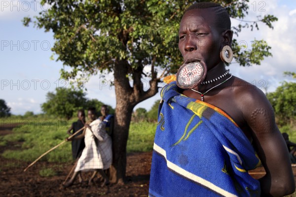 Mursi woman with lip plate in front of a tree in Mago National Park. A village can be seen in the