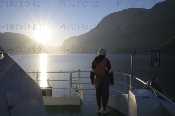 Person standing alone on a ferry at sunset with view of fjord and mountains, view from Columbus