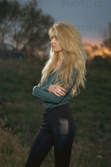 A woman stands with her arms crossed on a grassy hill. She faces the horizon where the sun is