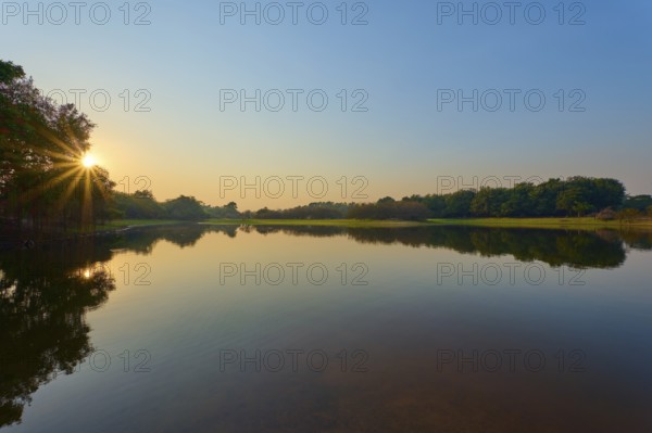 The first rays of sunshine fall on the calm lake, surrounded by green trees and clear skies,