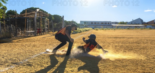 Two baseball players on a sunny field, one slides into a dusty base in a cloud of dirt while