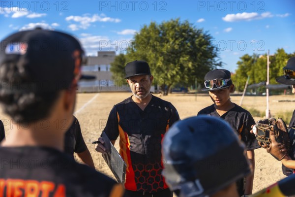 Baseball coach in a black and orange jersey and cap huddling with his youth team, discussing game