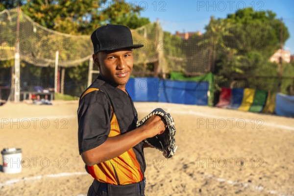 Confident young baseball player stands on dusty field gripping his glove, smiling at the camera and