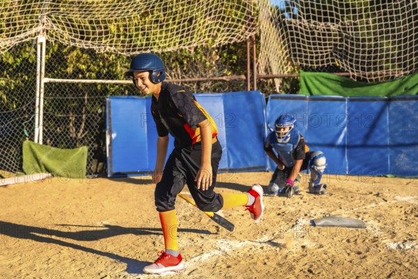 Young boy smiles while running on the dirt field, holding a bat and wearing a helmet and uniform,