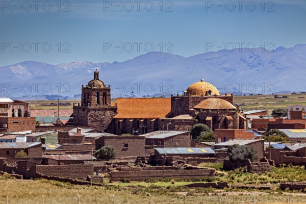 Historic church in a village with brick red roofs against a mountain backdrop under clear skies,