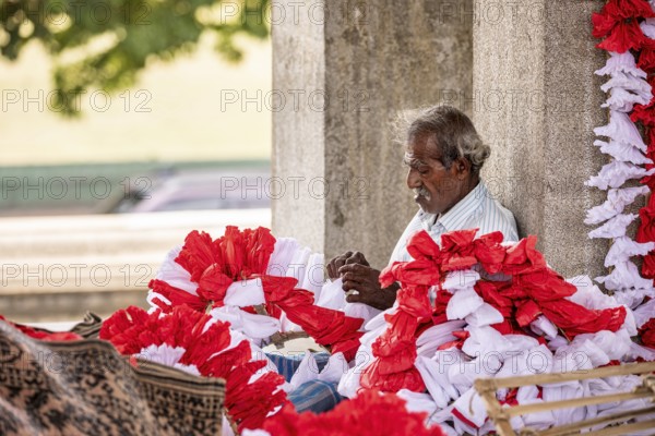 Elderly man arranges red and white flowers under a stone pillar in a warm, calm atmosphere, people
