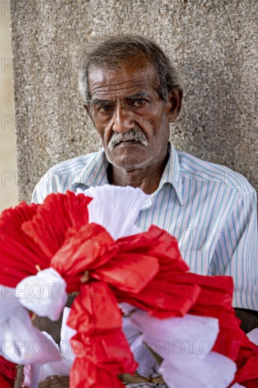 Close-up of an elderly man with red and white decorative flowers in the foreground, people making