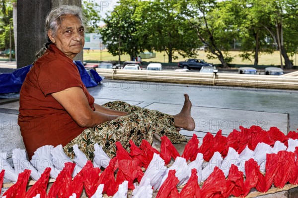 Elderly woman sitting outdoors working on red and white decorations, people making garlands in