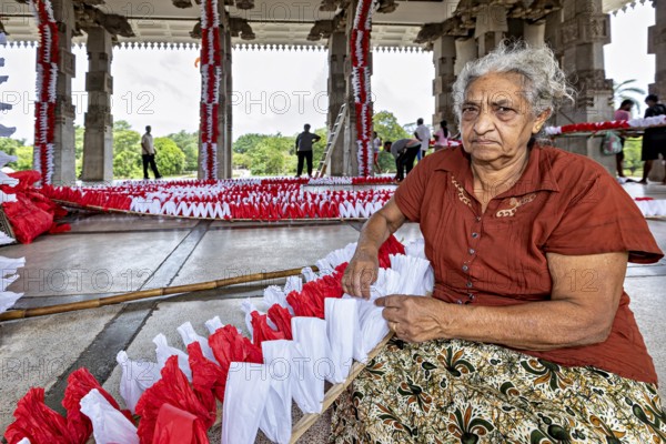 Elderly woman sitting and making red and white decorations in a hall, people making garlands in