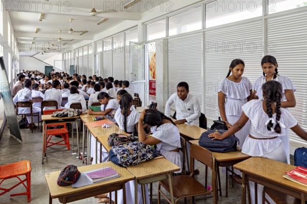 Students in uniforms sit in a bright classroom, some talk while others study, young people in a