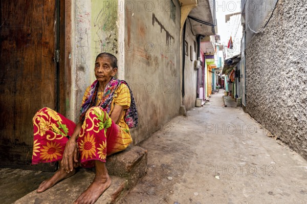Elderly woman sitting pensively in a narrow alley, people in the streets of Slave Island in Colombo