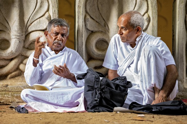 Two men in white robes are engaged in discussing in front of an artfully decorated temple, praying
