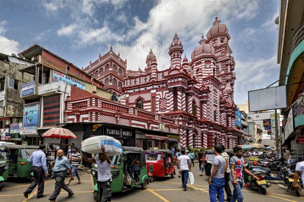 Bustling street scene with rickshaws and passers-by in front of a red architectural building, the