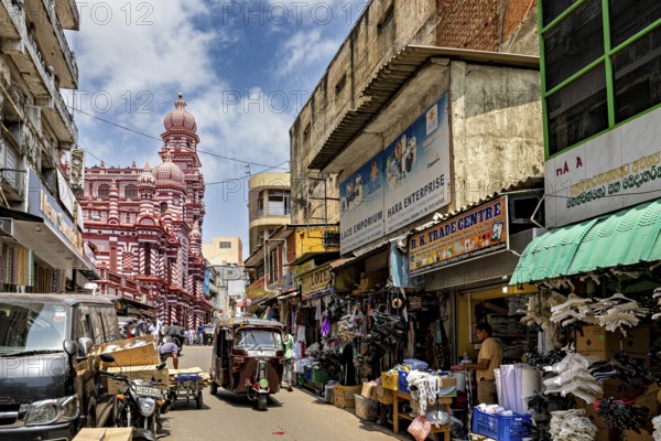 Bustling market street with people and stalls, red architectural building in the background, the