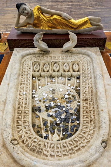 A reclining Buddha statue with gold robes above a carved stone surface filled with coins,