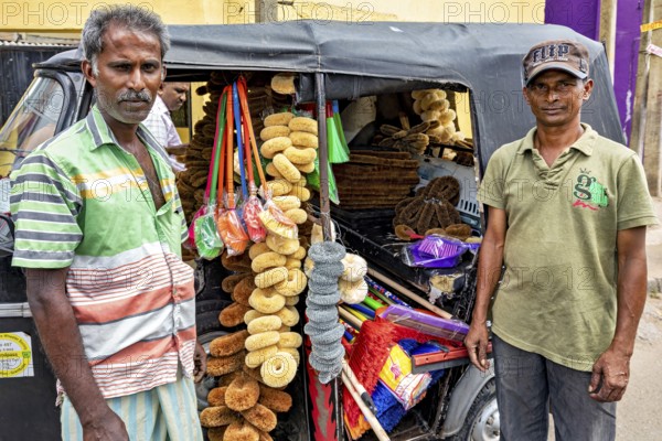 Two men stand in front of a tuk-tuk sales stand stocked with sponges on a busy street, tuk tuk as a
