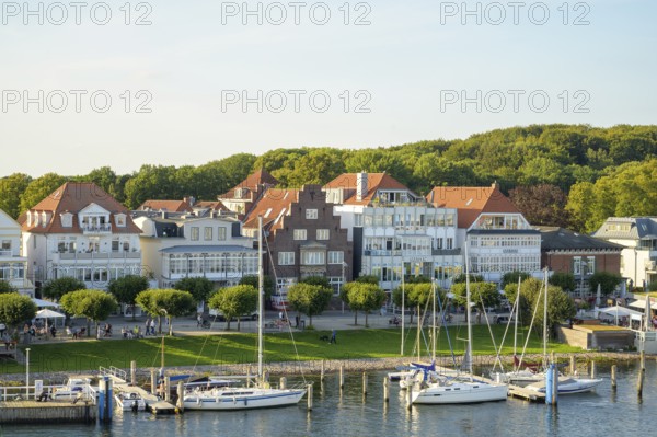 Picturesque houses and boats stand on a sunny harbor against a green forest backdrop,
