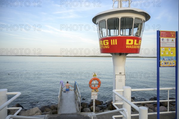 A modern DLRG watchtower looks out at the vast water while a bather sits on the jetty,