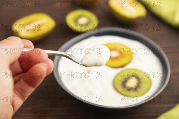 Yogurt, with kiwi in blue bowl on brown wooden background and green linen textile with hand, side