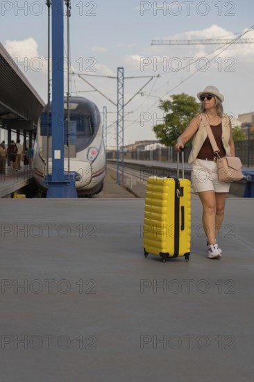 Middle aged woman pulling a bright yellow suitcase on a modern urban train platform, enjoying a