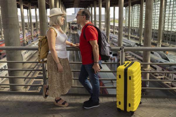 Happy mature tourist couple standing inside a large railway station, enjoying their journey with