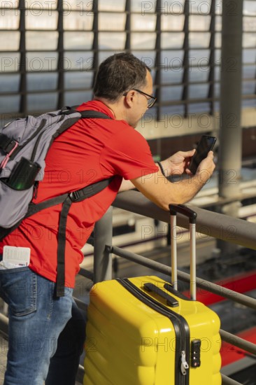 Mature male traveler using a smartphone at a train station while standing next to a yellow suitcase