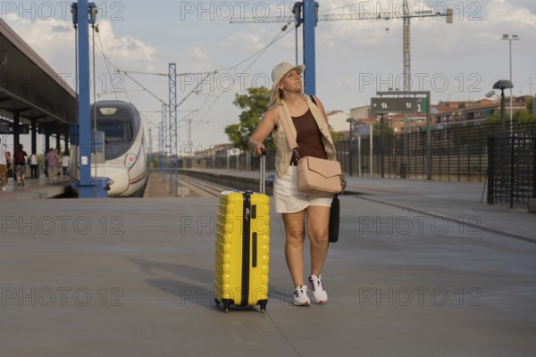 Adult woman walking along a modern train station platform, pulling a yellow suitcase while waiting