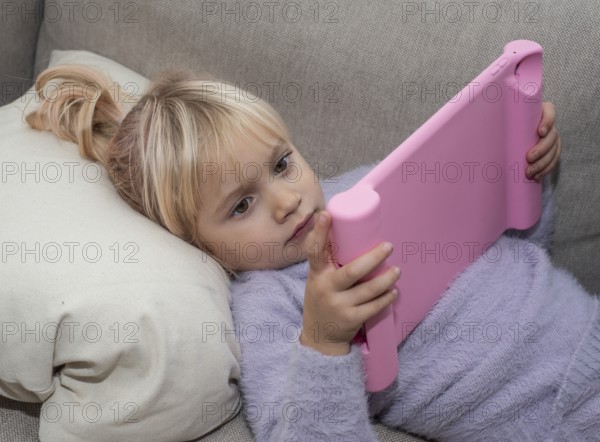 Portrait of a blonde girl, 4 years old, lying down reading a tablet in Ystad, Skåne County, Sweden,