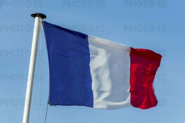 A French flag flutters in the wind on a white flagpole. The background is a clear, azure sky