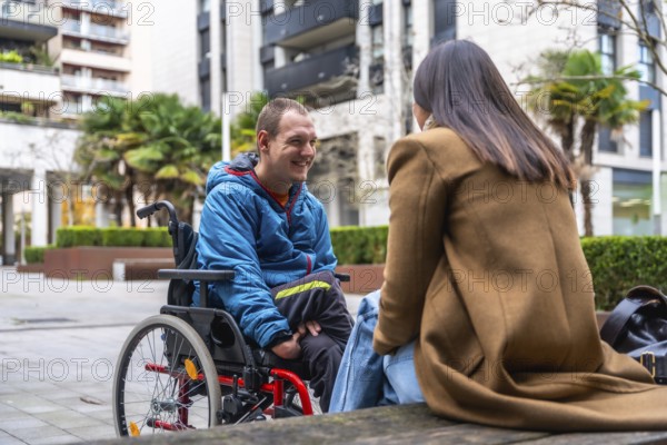 Young man in a wheelchair smiling and talking with a woman outdoors in an urban setting,