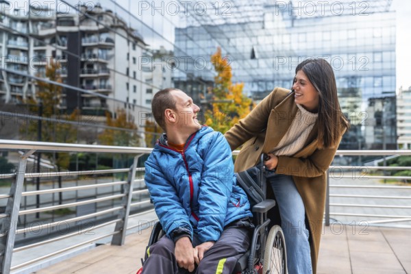 Caregiver woman pushes smiling young man in wheelchair along a city sidewalk, sharing a warm moment