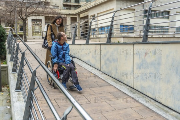 Woman pushing a young man with a disability in a wheelchair, navigating an accessible ramp with