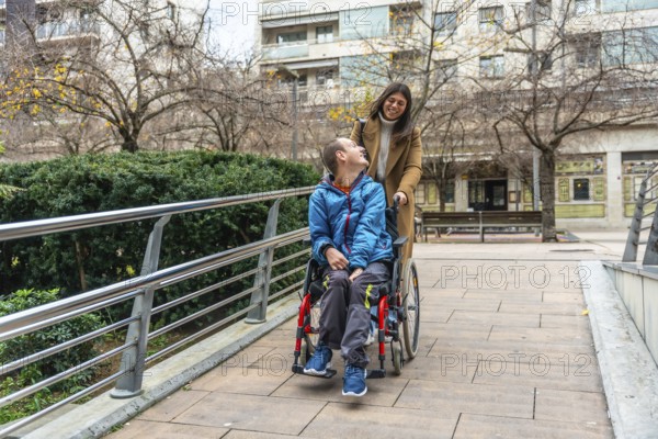 Caretaker pushing young man in wheelchair along accessible urban path, both smiling and sharing a